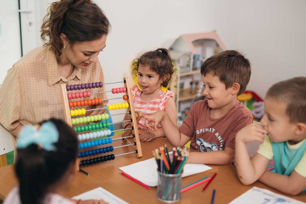 Educator kneeling beside two young children during cooperative block play in a bright classroom.